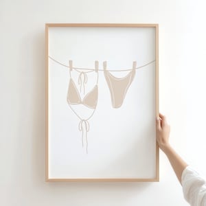 May include: A framed print depicting a beige bikini top and bottom suspended from a clothesline. The minimalist illustration is set against a white backdrop, within a light brown wooden frame. The artwork suggests a beach or summer theme.