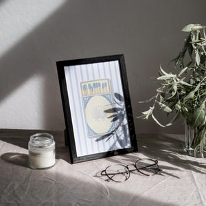 May include: A black framed print with a blue and white striped background. The print features a vintage illustration of a matchbox with blue matches and a seashell design.