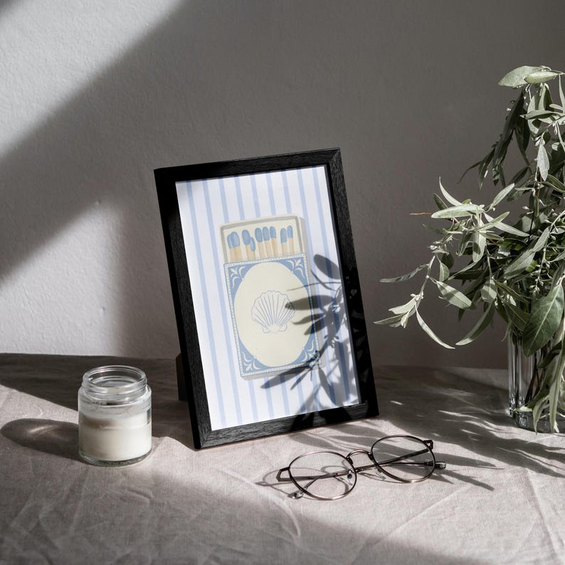 May include: A black framed print with a blue and white striped background. The print features a vintage illustration of a matchbox with blue matches and a seashell design.