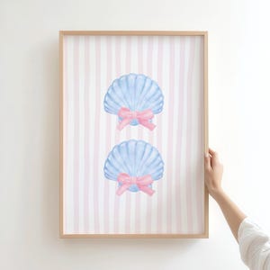 Op de afbeelding: Een ingelijste print met twee lichtblauwe schelpenillustraties, elk versierd met een roze strik, tegen een achtergrond van roze en witte verticale strepen. Het kunstwerk zit in een houten lijst.