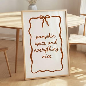 Könnte beinhalten: Ein rechteckiger Kunstdruck mit hellem Holzrahmen. Der weiße Druck zeigt einen braunen, wellenförmigen Rand mit einer Schleife oben. Der Text in der Mitte lautet "pumpkin spice and everything nice" in einer braunen, handschriftlichen Schriftart.