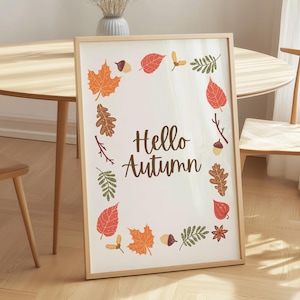 May include: A rectangular framed print with a light wood border featuring the text Hello Autumn in brown script. The design is surrounded by a border of colorful illustrated autumn leaves, acorns, and seeds in shades of orange, red, and green.