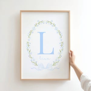 以下が含まれることがあります： 花輪の中に水色の文字「L」が入った額入りプリント。名前の「Lisa」は筆記体で下に書かれており、水色のリボンが付いています。フレームは明るい木の色です。