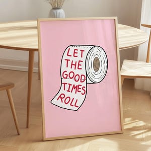 May include: A framed pink print featuring a cartoon illustration of a toilet paper roll with the words "LET THE GOOD TIMES ROLL" in red. The print is leaning against a wall, with a wooden table and chair in the background.