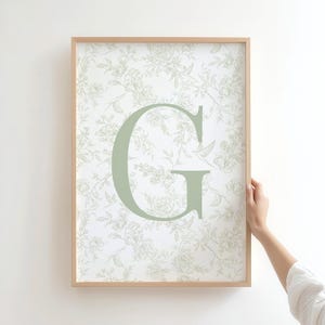 Op de afbeelding: Een ingelijste print met de letter "G" in een zachte groene tint. De achtergrond toont een delicaat bloemenpatroon in lichtgroen en wit. De houten lijst voegt een natuurlijke toets toe aan deze wanddecoratie.