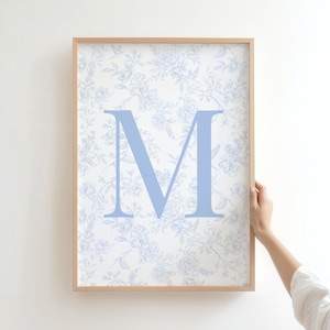 May include: Framed wall art featuring a light blue letter "M" on a floral patterned background. The artwork is in a light brown wooden frame. The floral pattern is a light blue and white design.