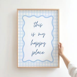 May include: A framed print with the words "this is my happy place" in a handwritten font. The print has a light blue gingham border and a scalloped edge. The frame is made of light-colored wood.