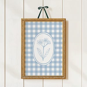 May include: A framed floral print with a light blue and white gingham pattern. The gold-coloured frame surrounds a print with an oval floral design. A dark green ribbon is used for hanging the framed artwork. The print is a decorative item.