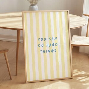 Puede incluir: Una pieza de arte mural rectangular enmarcada con rayas verticales de color amarillo pálido y blanco. El centro de la impresión muestra el texto azul "YOU CAN DO HARD THINGS" en una fuente sencilla sin remates. El marco es de madera clara.
