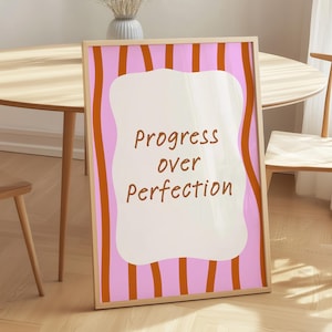 Op de afbeelding: Een ingelijste kunstafdruk met de woorden "Progress over Perfection" in bruin schrift. Het kunstwerk toont een witte, golvende vorm tegen een roze achtergrond met verticale oranje strepen. Het frame heeft een lichte houtkleur.