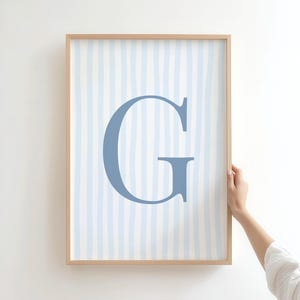 Pode incluir: Uma impressão emoldurada com uma grande letra "G" azul claro sobre um fundo de listras verticais azul claro. A obra de arte está em uma moldura de madeira clara, segurada por uma mão.