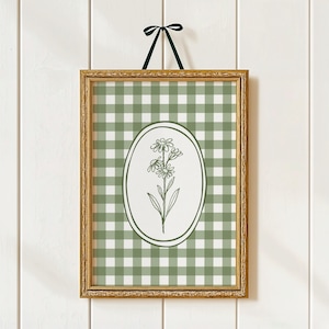 May include: A framed print with a green and white gingham pattern. An oval in the centre displays a floral line drawing. The gold frame is suspended by a green ribbon. The artwork is mounted on a white wooden wall.