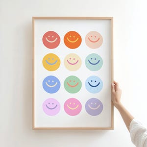 May include: A framed print featuring a grid of twelve colorful smiley faces. Each smiley face is a different color, including red, orange, yellow, blue, green, and purple. The print has a light wood frame and is displayed against a white wall.