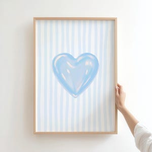 May include: A framed art print featuring a light blue heart on a background of vertical light blue stripes. The artwork is in a wooden frame. The image is held by a person.