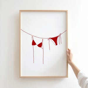 Peut inclure: Une impression d'art encadrée présentant une illustration minimaliste d'un bikini rouge suspendu à une corde à linge. L'œuvre est posée sur un fond blanc, avec une esthétique simple et épurée. Le cadre est de couleur bois clair.