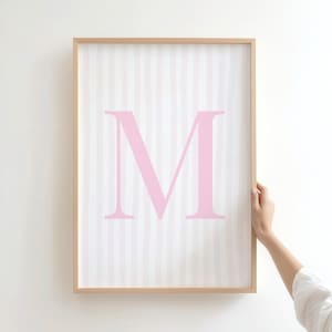 May include: A framed print featuring a large pink letter "M" centered on a white background with vertical pink stripes. The frame is a light wood color. The print is held by a person's hand.