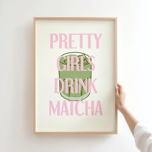 May include: A framed art print with the text "PRETTY GIRLS DRINK MATCHA" in pink, with a green matcha drink illustration. The artwork is in a light wood frame, held by a person's hand.