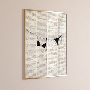 May include: A framed art print with a gold frame, showing a minimalist line drawing of two black bikinis hanging on a washing line. The background is a collage of old newspaper pages.