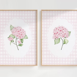 Op de afbeelding: Twee ingelijste bloemenprints. Elk heeft een aquarel hortensia in roze en groen, geplaatst op een witte geschulpte ovaal op een roze ruitjesachtergrond. De frames zijn van licht hout.