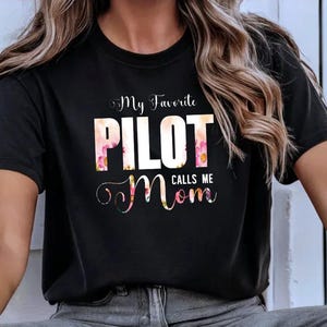 Peut inclure: T-shirt noir avec un texte blanc qui dit "My Favorite Pilot Calls Me Mom". Le texte est entouré d'un motif floral.