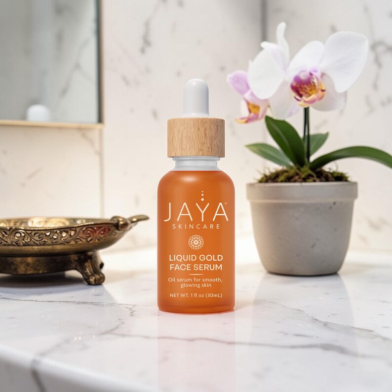 May include: An orange glass bottle of Jaya Skincare Liquid Gold Face Serum with a wooden cap and dropper. The label reads "Oil serum for smooth, glowing skin" and "NET WT. 1 fl oz (30mL)". The bottle sits on a marble surface near a decorative dish and a potted orchid.