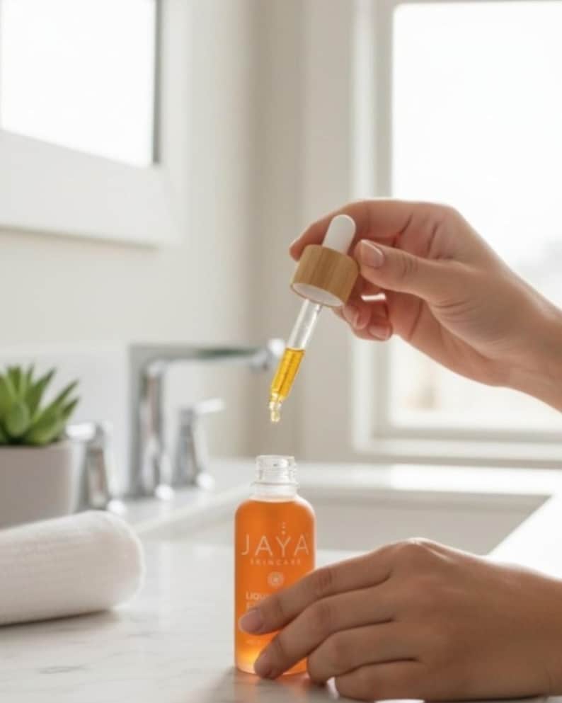 May include: A clear glass bottle of Jaya Skincare liquid serum with a dropper. The serum is amber-colored. The bottle is held by a person's hands. The dropper is made of glass and wood. The Jaya Skincare logo is visible on the bottle.