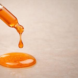 May include: A close-up shot of a glass dropper dispensing a golden-orange liquid. A single drop is falling, and a small pool of the liquid has already formed on the surface. The dropper is angled downward, and the background is a neutral beige.