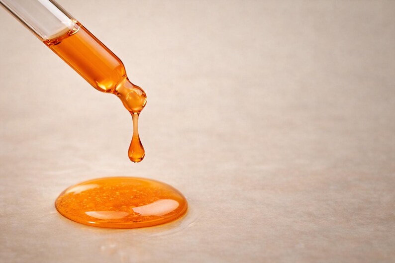 May include: A close-up shot of a glass dropper dispensing a golden-orange liquid. A single drop is falling, and a small pool of the liquid has already formed on the surface. The dropper is angled downward, and the background is a neutral beige.