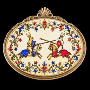 May include: An oval-shaped brooch featuring a gold-toned metal frame with a shell-like top detail. The centre depicts two rabbits jousting on horseback, one brown and one white, surrounded by a colourful floral border in red, blue, and green.
