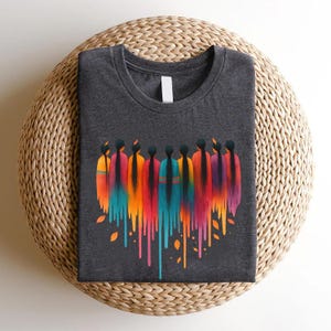 May include: Dark gray t-shirt featuring a vibrant graphic of figures with long black hair and colorful, flowing garments in shades of orange, yellow, blue, and purple. The design includes dripping paint effects and autumn leaves.
