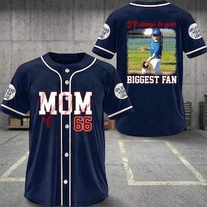 以下が含まれることがあります： 白のトリムとボタンが付いた、濃いネイビーブルーの野球ジャージ。前面には、赤い野球のステッチと「66」が赤で「MOM」が白で表示されています。背面には写真と「I'll always be your BIGGEST FAN」というテキストが表示されています。