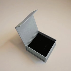 May include: An open, gray jewelry box with a black velvet interior. The box is rectangular with a hinged lid, and the interior has slots for holding jewelry. The box is sitting on a light-colored surface.