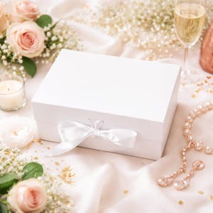 May include: A white gift box with a satin ribbon bow, surrounded by roses, baby's breath, a lit candle, a champagne glass, and a necklace with earrings. The scene is set on a white satin cloth.