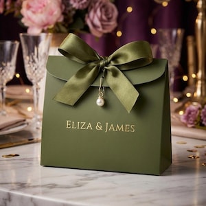 May include: An olive green gift bag with a satin ribbon bow in the same color. A small gold-toned safety pin with a white pearl charm is attached to the center of the bow. The text ELIZA & JAMES is printed in gold lettering on the front of the bag.