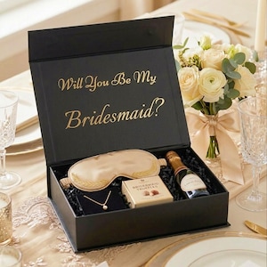May include: A black gift box with the gold text Will You Be My Bridesmaid? containing a champagne mini bottle, a gold sleep mask with embroidered leaf details, a small box of bridesmaid truffles, and a delicate gold necklace with a pearl pendant.
