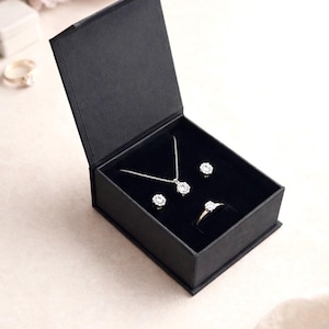 May include: A black jewelry box with an open lid reveals a necklace, earrings, and a ring. The jewelry features clear, round gemstones. The box is on a light-colored surface, with flowers in the background.
