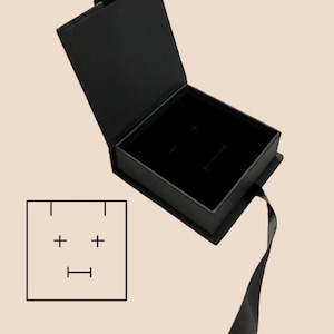 May include: Black jewelry box with a hinged lid, open to reveal a black velvet interior. A black ribbon extends from the box. A small square graphic with a face-like design is in the lower left corner.
