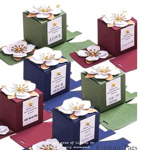 May include: An assortment of elegant candy boxes in various colors, including green, burgundy, and navy blue. Each box is adorned with a white paper flower and a small tag. The text on the boxes reads "Thank you for celebrating with us".