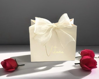 Könnte beinhalten: Elfenbeinfarbene Geschenktüte mit einer durchsichtigen Schleife und goldenem Text mit der Aufschrift "Victoria". Zwei rote Rosen liegen auf der Oberfläche. Die Tüte steht auf einer weißen Oberfläche mit grauem Hintergrund. Die Tüte ist eine Geschenktüte.