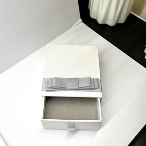 May include: A white gift box with a gray bow and a pull-out drawer. The box is open, revealing a gray interior. The bow is made of a textured ribbon. The box is sitting on a white surface.