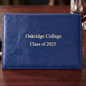 May include: A rectangular navy blue diploma cover with a textured finish and stitched edges. The front features gold foil lettering that reads Oakridge College and Class of 2025 centered in the middle of the cover in a serif font style.