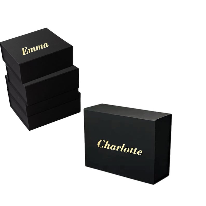 May include: Black gift boxes stacked and arranged, with the name "Emma" in gold lettering on the top box. Another box with the name "Charlotte" in gold lettering is also visible. The boxes are likely for jewelry or small gifts.