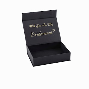 May include: Black gift box with a hinged lid, open to reveal an empty interior. The box features gold script text on the inside of the lid that reads, "Will You Be My Bridesmaid?" The box is set against a white background.
