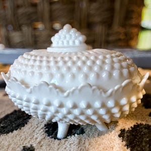 Puede incluir: Un plato blanco cubierto de hobnail con tapa. El plato tiene un borde festoneado y se apoya en tres patas pequeñas. La tapa está rematada con un pequeño pomo decorativo. El plato es probablemente de cerámica o porcelana.