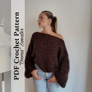 Könnte beinhalten: Ein brauner Off-Shoulder-Pullover mit langen Ärmeln. Der Pullover ist aus einem strukturierten Garn gefertigt und wird mit einer blauen Jeans kombiniert. Der Text auf der linken Seite lautet "PDF Crochet Pattern 'Emma' Sweater".