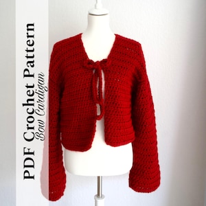 Könnte beinhalten: Ein roter Häkelcardigan mit Schleifenverschluss. Der Cardigan hat lange Ärmel und eine verkürzte Länge. Der Text "PDF Crochet Pattern Bow Cardigan" ist im Bild sichtbar.