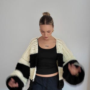 Pode incluir: Um cardigan de croch&ecirc; feito &agrave; m&atilde;o, com listras creme e pretas. As mangas s&atilde;o largas e em forma de sino. O cardigan &eacute; usado sobre uma regata preta e cal&ccedil;as azul-escuras. A palavra "chap" &eacute; vis&iacute;vel no peito.