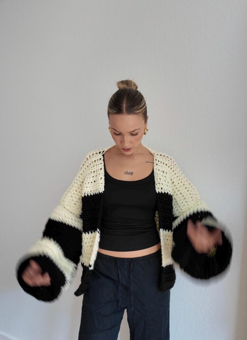 Pode incluir: Um cardigan de croch&ecirc; feito &agrave; m&atilde;o, com listras creme e pretas. As mangas s&atilde;o largas e em forma de sino. O cardigan &eacute; usado sobre uma regata preta e cal&ccedil;as azul-escuras. A palavra "chap" &eacute; vis&iacute;vel no peito.