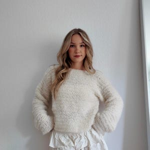 May include: Cream-colored, hand-knitted sweater with a crew neck and long sleeves. Paired with a white, ruffled mini skirt. The sweater has a textured, fluffy appearance. The outfit is casual and stylish.