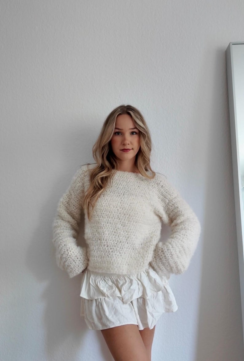 May include: Cream-colored, hand-knitted sweater with a crew neck and long sleeves. Paired with a white, ruffled mini skirt. The sweater has a textured, fluffy appearance. The outfit is casual and stylish.